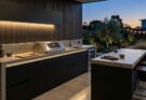 Modern minimalist outdoor kitchen with polished concrete, matte black cabinets, stainless steel grill, and warm LED lighting.